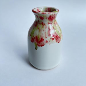 May include: A small, white ceramic vase with a rounded body and a flared neck. The top portion is decorated with a vibrant pattern of red and green spots against a white background. The vase is a decorative item.