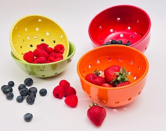 Op de afbeelding: Een verzameling kleurrijke keramische kommen met drainagegaten, gevuld met vers fruit. De kommen zijn geel, groen, oranje en rood en bevatten frambozen, aardbeien en blauwe bessen. Een levendige presentatie van fruit.