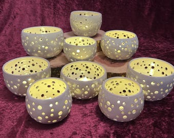 May include: A collection of ceramic candle holders in various sizes. Each holder is light gray with numerous small, evenly spaced holes. The interiors are yellow, and some hold lit candles, creating a warm glow. The holders are arranged on a wooden surface.