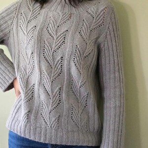 Puede incluir: Un suéter de punto gris claro con un patrón de hojas. El suéter tiene un cuello redondo y mangas largas.