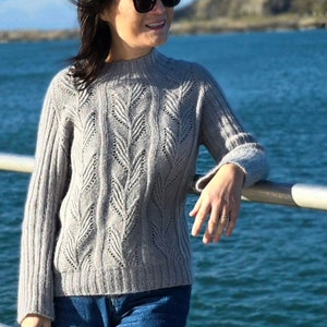 Puede incluir: Una mujer lleva un suéter de punto gris claro con un patrón de punto de cable y cuello alto. Lleva pantalones vaqueros azules y gafas de sol.