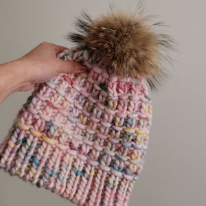 Op de afbeelding: Een roze gebreide beanie met een veelkleurig garen en een bruine nep bont pompon.