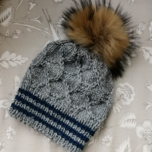 Op de afbeelding: Grijze gebreide beanie met een blauwe en grijze gestreepte band en een bruine nep bont pompon.