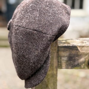 Puede incluir: Gorra de estilo newsboy de tweed marrón con una corona redondeada y una visera corta y rígida. La gorra está hecha de una tela texturizada y descansa sobre un poste de valla de madera. Tiene un botón lateral.