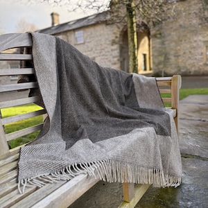 May include: A gray and brown herringbone-patterned blanket with fringe detail draped over a wooden bench. The blanket features a color block design with a lighter gray section. The scene is outdoors.