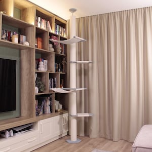 Könnte beinhalten: Ein weißer Kratzbaum mit mehreren Plattformen und einer zentralen Säule. Die Plattformen sind rechteckig und mit einem hellfarbigen Stoff bedeckt. Der Kratzbaum befindet sich in einem Raum mit einem Bücherregal und einem beigen Vorhang.