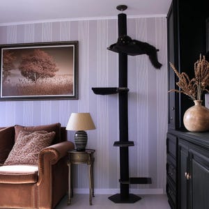 May include: A black cat tree with multiple platforms and a scratching post. The cat tree is in a living room with a brown sofa, a framed picture, and a lamp on a side table. A black cat is resting on the top platform.