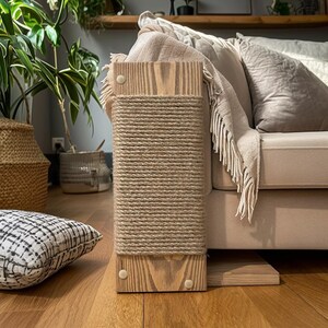 May include: A natural wood cat scratching post with a sisal rope scratching surface. The post is rectangular and has a wooden base and top. The sisal rope is tightly wrapped around the center section. The post is next to a beige sofa.