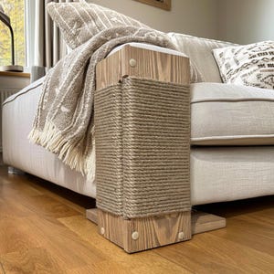 May include: A wooden cat scratching post with a natural rope wrapping. The post is rectangular and sits next to a beige sofa. A beige throw blanket with fringe and a patterned pillow are on the sofa.