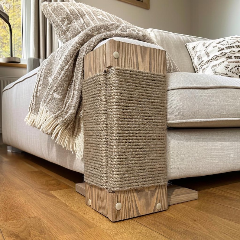 May include: A wooden cat scratching post with a natural rope wrapping. The post is rectangular and sits next to a beige sofa. A beige throw blanket with fringe and a patterned pillow are on the sofa.
