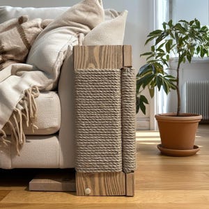 Puede incluir: Un rascador de gato de madera con un envoltorio de cuerda natural. El poste es rectangular y tiene un acabado de madera clara. La cuerda es de color beige neutro y el poste se coloca junto a un sofá beige. Una planta en maceta está en el fondo.