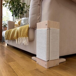 May include: A wooden cat scratching post with a square base and top. The post is wrapped in white rope. The post is next to a beige sofa with a yellow blanket and pillows. The floor is made of wood.