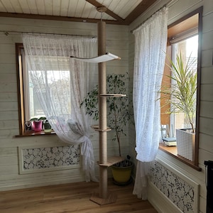 May include: A tall, natural-coloured cat tree with multiple platforms and a sisal-covered scratching post. The tree is in a room with white walls and lace curtains. A potted plant is next to the tree.