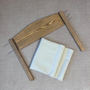 May include: A wooden frame with two sets of hooks on each side and a beige fabric cushion. The frame is made of light brown wood with a natural wood grain.