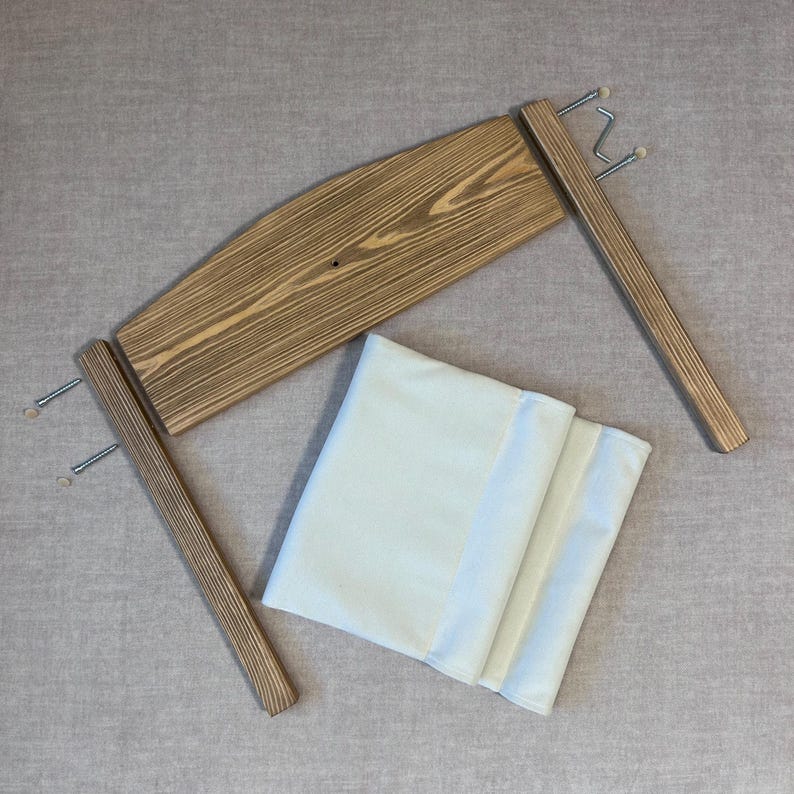 May include: A wooden frame with two sets of hooks on each side and a beige fabric cushion. The frame is made of light brown wood with a natural wood grain.