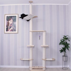 May include: A multi-level cat tree with a black cat resting on a hammock-style platform. The cat tree features multiple platforms, scratching posts, and a hanging toy. The structure is made of light-colored wood and beige fabric, set against a striped wall.