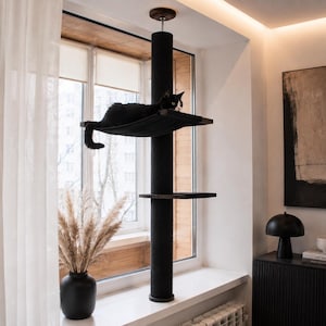 Pode incluir: Uma árvore para gatos do chão ao teto com um poste central coberto de carpete preto, uma plataforma de rede de tecido preto com um gato preto e branco a descansar, e uma plataforma preta mais pequena, fixadas entre o chão e o teto.