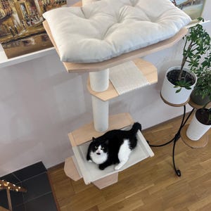 May include: A white cat with black markings is relaxing on a beige hammock attached to a wooden cat tree. The cat tree has a beige cushion on top and a beige sisal scratching post. The cat tree is against a white wall.