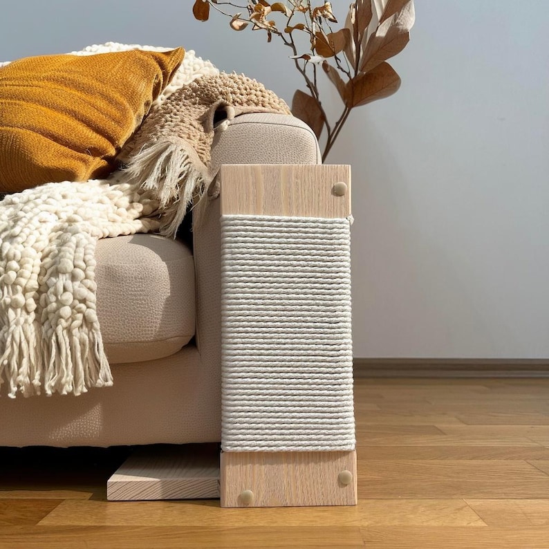 May include: A wooden cat scratching post with a natural finish and a wrapped sisal rope scratching surface. The post is rectangular and has a modern design, perfect for feline entertainment and claw care. The post is placed next to a beige sofa.