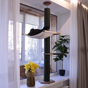 May include: A black cat lounging on a hammock-style bed attached to a tall, green cat tree. The cat tree has a wooden base and a platform below the hammock. The cat tree is positioned in front of a window with white curtains.