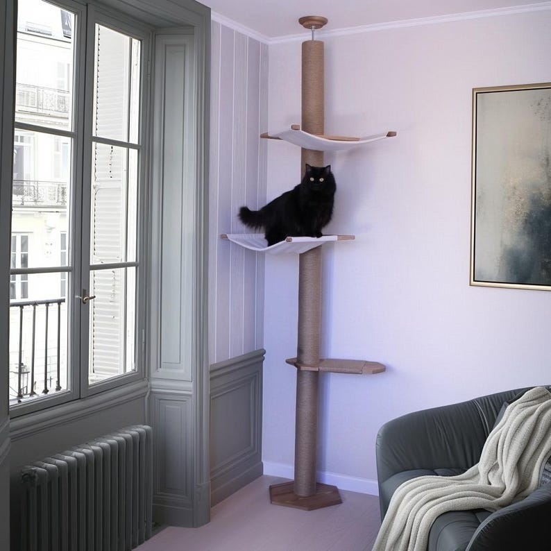 May include: A tall cat tree with multiple platforms and a central scratching post. A black cat sits on one of the white fabric platforms. The tree is made of wood and rope, and is in a room with a window and a painting.