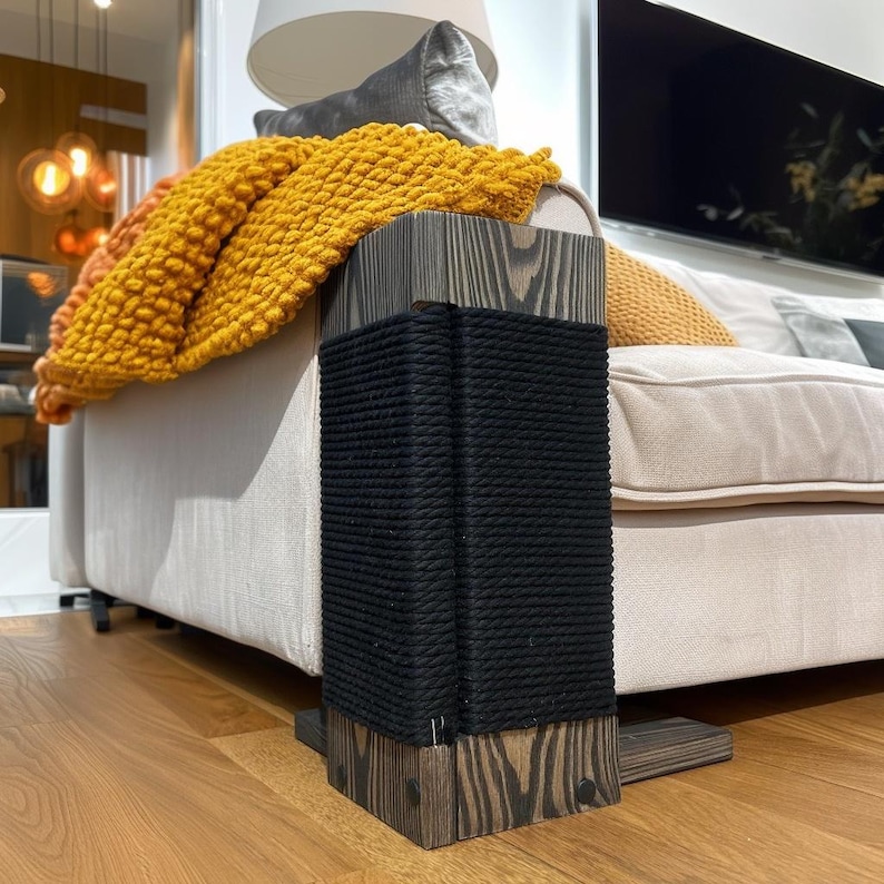May include: A wooden cat scratching post with a dark gray finish and black rope wrapping. The post is rectangular and sits on a wooden base. A yellow blanket is draped over a beige sofa in the background.