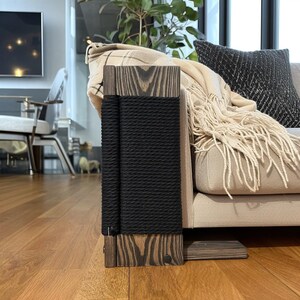 May include: A wooden cat scratching post with a black rope scratching surface. The post is made of dark wood with a natural grain pattern. The post is next to a beige sofa with a fringed blanket and a black pillow.