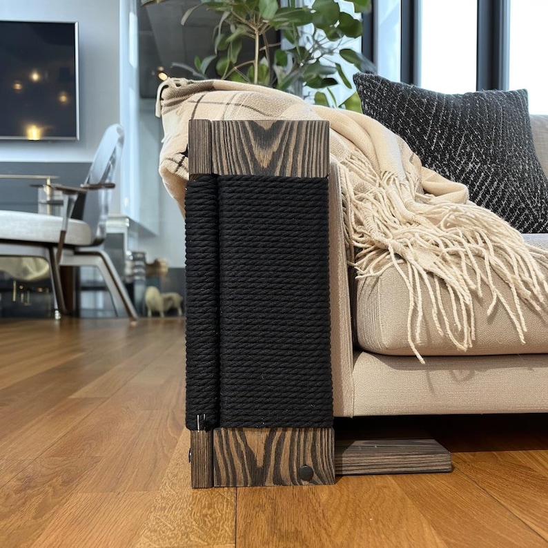 May include: A wooden cat scratching post with a black rope scratching surface. The post is made of dark wood with a natural grain pattern. The post is next to a beige sofa with a fringed blanket and a black pillow.