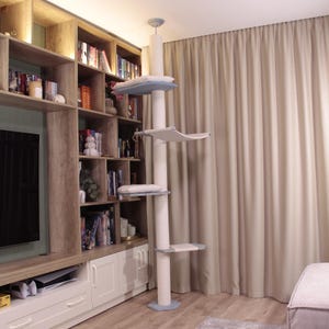 Könnte beinhalten: Ein hoher, weißer Kratzbaum mit mehreren Ebenen, einschließlich Plattformen und einer Hängematte. Der Kratzbaum hat hellblaue Akzente und steht in einem Wohnzimmer mit Bücherregal und Vorhängen.