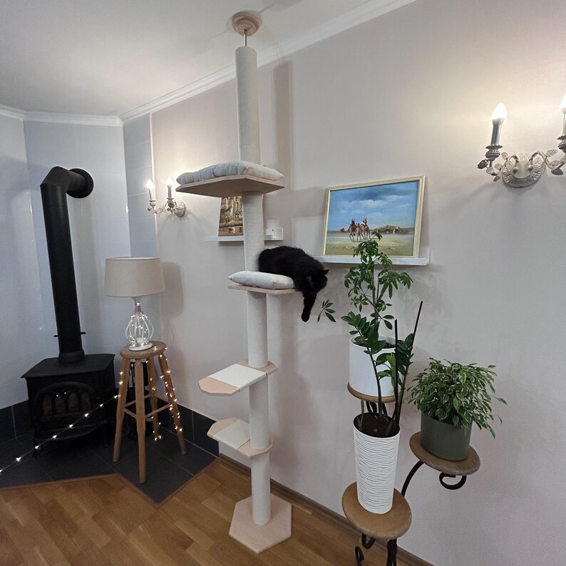May include: A beige cat tree with multiple platforms and a sisal-covered scratching post. A black cat is lounging on one of the platforms. The cat tree is in a room with a fireplace, a lamp, and plants.