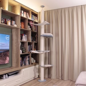 Op de afbeelding: Een hoge, witte kattenboom met meerdere niveaus en platforms. De kattenboom heeft een centrale kolom met een reeks platforms en rustplaatsen, waaronder een hangmat en een kussenbed. Het ontwerp is modern en past goed in het interieur.