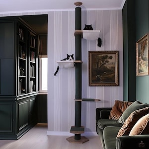 May include: A dark green cat tree with multiple platforms and a central scratching post. Two cats are resting on the platforms. The cat tree is in a room with a dark green bookcase, a painting, and a green sofa.