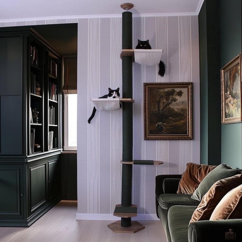 May include: A dark green cat tree with multiple platforms and a central scratching post. Two cats are resting on the platforms. The cat tree is in a room with a dark green bookcase, a painting, and a green sofa.