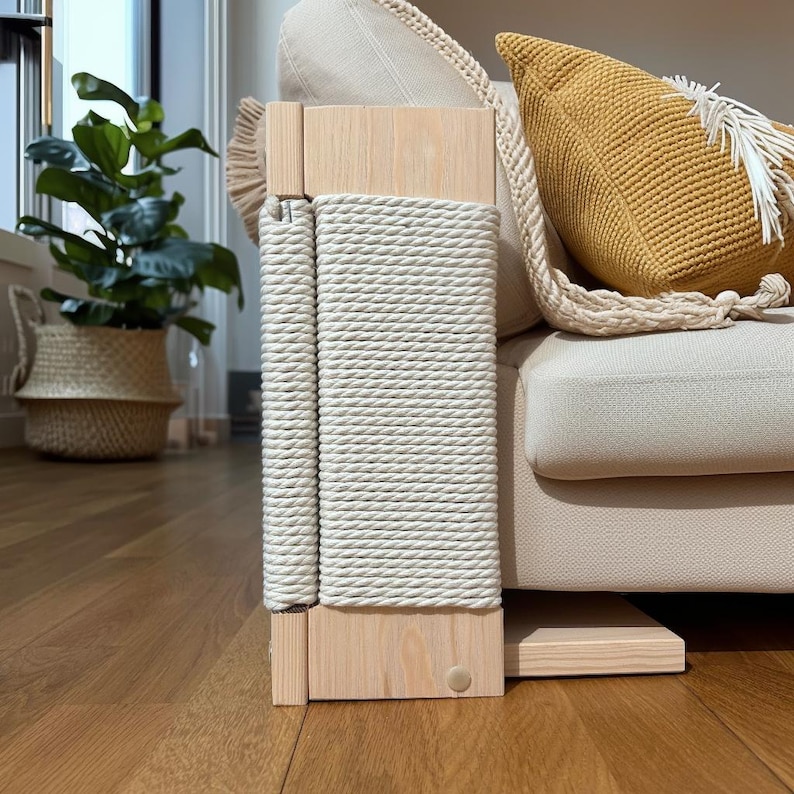 May include: A light-colored wooden cat scratching post with a cream-colored rope wrapped around it. The post is attached to a beige sofa. A yellow pillow with a white tassel is on the sofa.