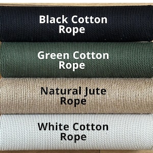 May include: Four spools of rope in different colors: black cotton, green cotton, natural jute, and white cotton. Each spool has text labeling the type of rope. The ropes are tightly wound and appear to be made of natural fibers.