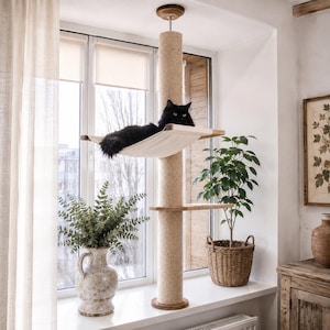 May include: A floor-to-ceiling cat tree featuring a tall, cylindrical scratching post wrapped in beige sisal rope. It includes a suspended off-white fabric hammock where a long-haired black cat is resting, and a small wooden platform attached.