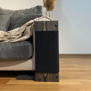 May include: A wooden cat scratching post with a black rope scratching surface. The post is attached to a sofa arm. The wood has a dark stain and is secured with black screws. A neutral-toned blanket and pillow are on the sofa.