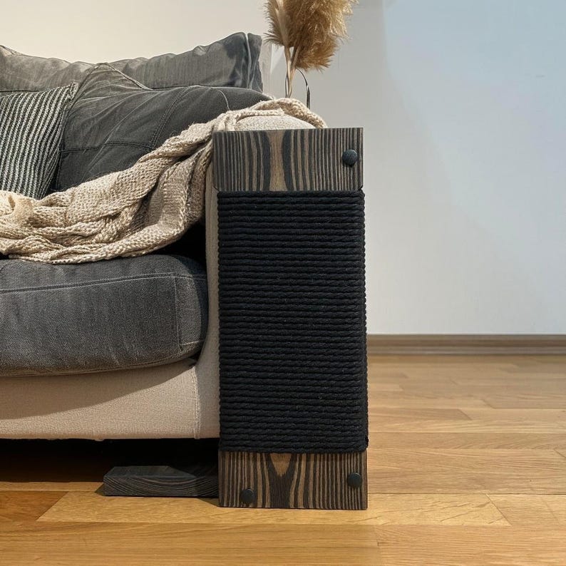 May include: A wooden cat scratching post with a black rope scratching surface. The post is attached to a sofa arm. The wood has a dark stain and is secured with black screws. A neutral-toned blanket and pillow are on the sofa.