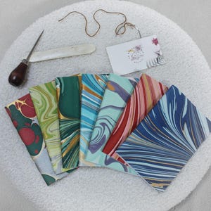 May include: A set of eight marbled paper notebooks with different color combinations. The notebooks are arranged in a fan shape, with the covers facing outward. The marbling patterns are unique and intricate, with swirls of blue, green, red, and gold.