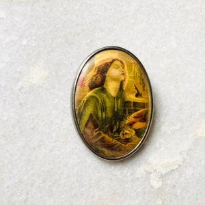 May include: Oval-shaped silver brooch with a vintage image of a woman in a green dress, with her eyes closed, looking up. The image is surrounded by a silver frame.