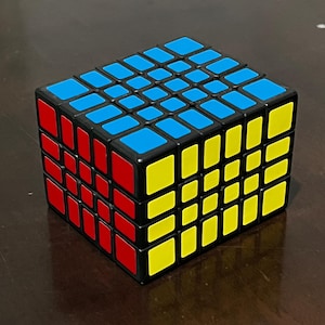 May include: A black 5x5 Rubik's Cube with blue, red, and yellow squares on the visible sides.