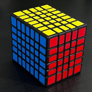Pode incluir: Um cubo de Rubik de seis camadas com uma moldura preta. As faces do cubo são compostas por pequenos quadrados amarelos, azuis e vermelhos. O cubo é um brinquedo de quebra-cabeça.