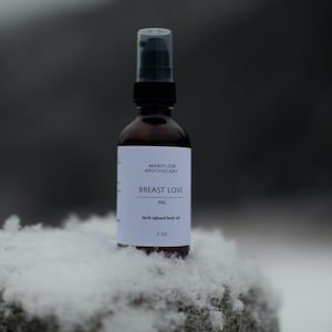 以下が含まれることがあります： 白いラベルに「MARILOW APOTHECARY BREAST LOVE OIL Herb infused body oil 2 OZ」と書かれた茶色のガラス瓶。瓶は白い表面の上に置かれている。