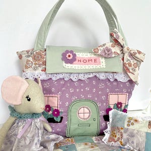 May include: A fabric house-shaped handbag in lavender with a green door, windows, and a floral top flap labelled HOME. Beside it sits a beige fabric mouse doll wearing a lavender floral dress and a small patchwork quilt with lace trim.
