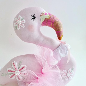 May include: A pink flamingo plush toy with a white lace collar and a pink tulle skirt. The flamingo has embroidered flowers and a black eye.