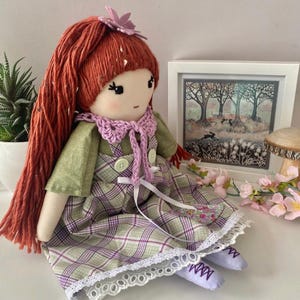 Puede incluir: Una muñeca hecha a mano con pelo de lana rojo, una camisa verde, una falda a cuadros con un borde de encaje blanco y zapatos morados. La muñeca lleva una bufanda de ganchillo rosa y tiene una flor rosa en el pelo.