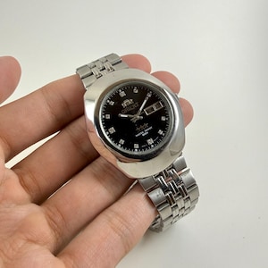 May include: A silver-toned Orient watch with a black face. The watch features a rectangular case, a linked metal band, and a date display. The watch face has small diamond-shaped markers and the brand name "Orient" is visible.