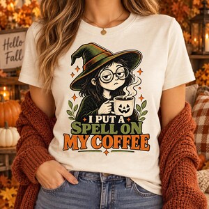 May include: A cream-coloured t-shirt featuring a graphic of a witch wearing a green hat and round glasses, holding a steaming mug with a jack-o'-lantern face. The text reads I PUT A SPELL ON MY COFFEE in green and orange block lettering.