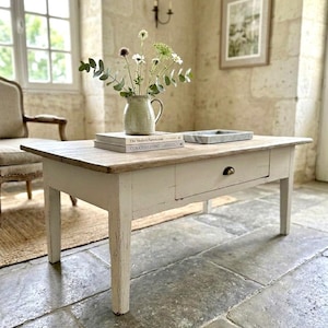 Peut inclure: Une table basse rectangulaire avec une base peinte en blanc vieilli et un plateau en bois naturel. La table comporte un tiroir unique avec une petite poignée en métal. Sur le dessus sont posés deux livres empilés et un pichet en céramique.