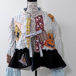 May include: A long-sleeve button-up shirt featuring a white grid pattern with various fabric patches including pumpkins, ghosts, and candy corn. The shirt has a black ruffled hem, a row of gold circular buttons, and light blue gingham cuffs.